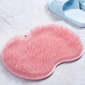 A pink silicone scrubber mat with dense bristles, shown on a light gray tiled floor next to a pair of light blue slippers. A pink label in the bottom.