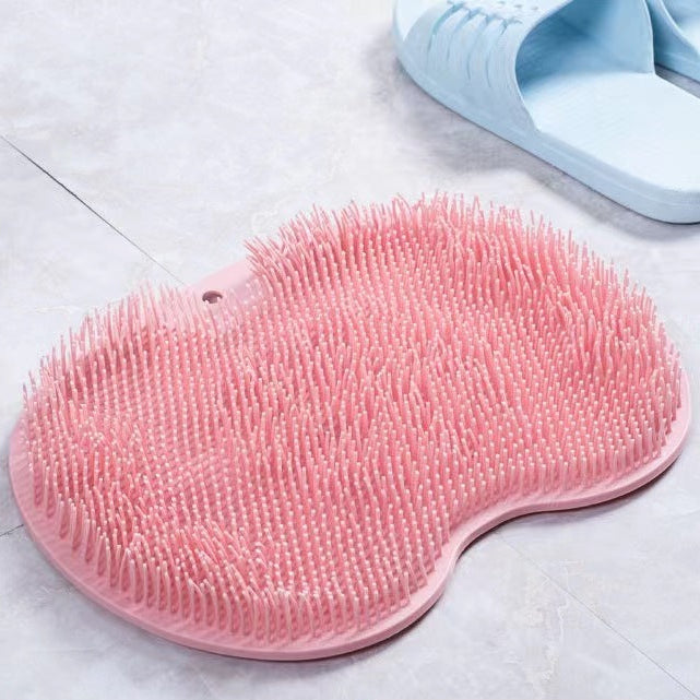 A pink silicone scrubber mat with dense bristles, shown on a light gray tiled floor next to a pair of light blue slippers. A pink label in the bottom.