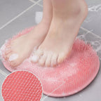 A pink foot scrubber with dense bristles is shown under a person's feet in soapy water on a tiled floor, with a circular inset close-up of the scrubber's bristle texture.