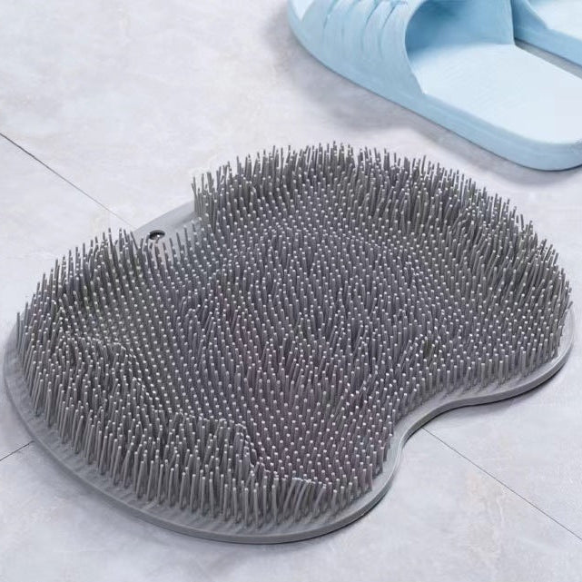 A gray silicone foot scrubber with dense bristles, shown on a light gray tiled floor next to a pair of light blue slippers. A black label with white.